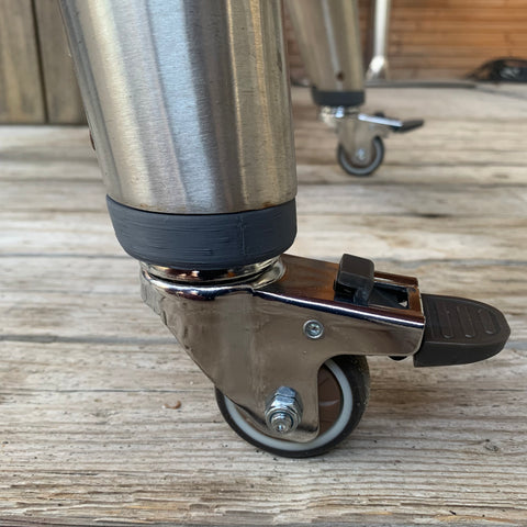 Grijze prototype wheelkit met zilveren wielen en rem, gemonteerd onder een metalen barbecuepoot op houten planken.