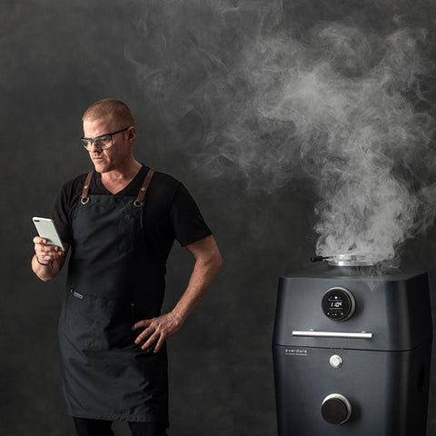 Man in schort bedient Everdure 4K Mat Zwart kamado grill met smartphone, rook stijgt op.